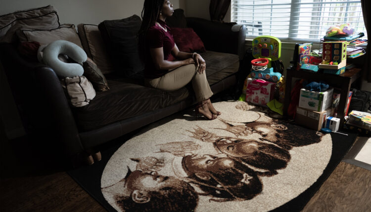 A photo of a Black woman sitting for a portrait indoors by a window. The carpet below her couch has an image of four young Black girls praying.