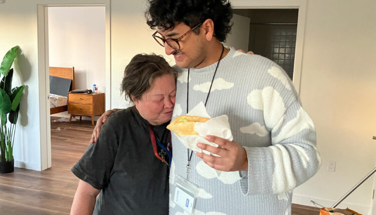 Julian Prado, a tall 29-year-old man with a nose piercing and black mustache, hugs Carla Brown while holding a taco in his other hand. Brown is a 60-year-old woman.